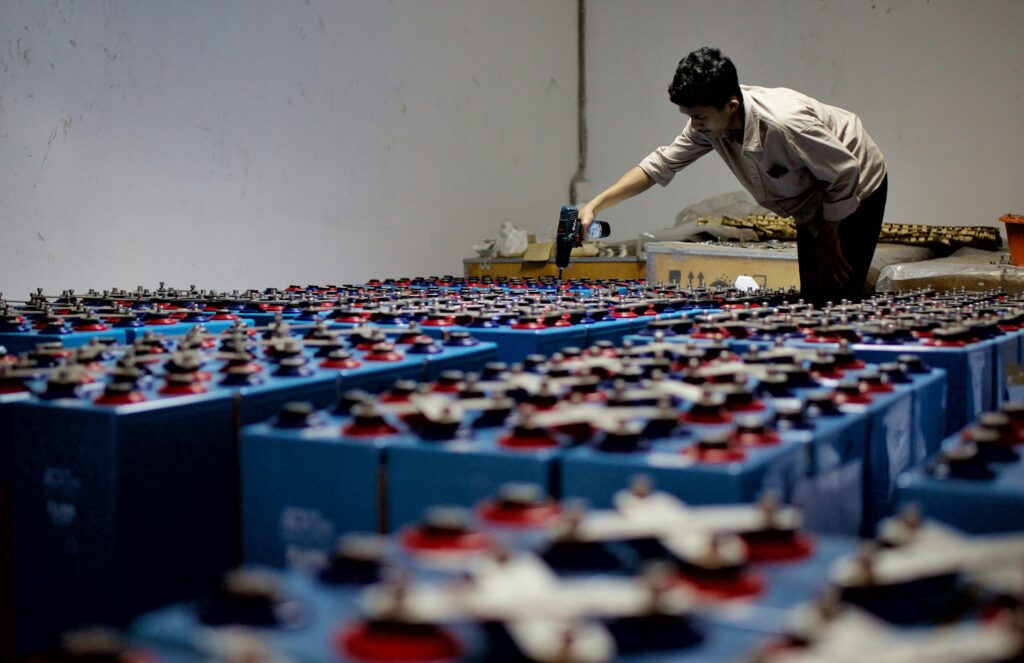 A worker checking many industrial batteries inside a facility. Indoor, industrial setting.